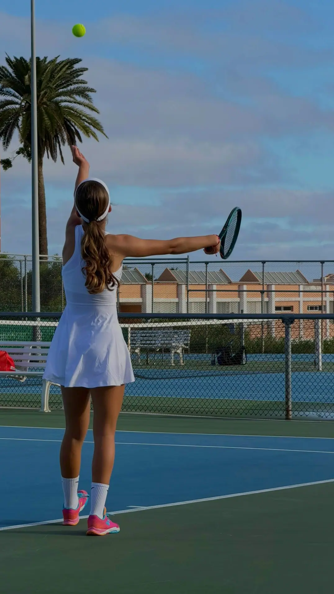 Nicole, de espaldas y con vestido blanco, lanza la bola para sacar en una pista azul; detrás, una palmera y un cielo con nubes.