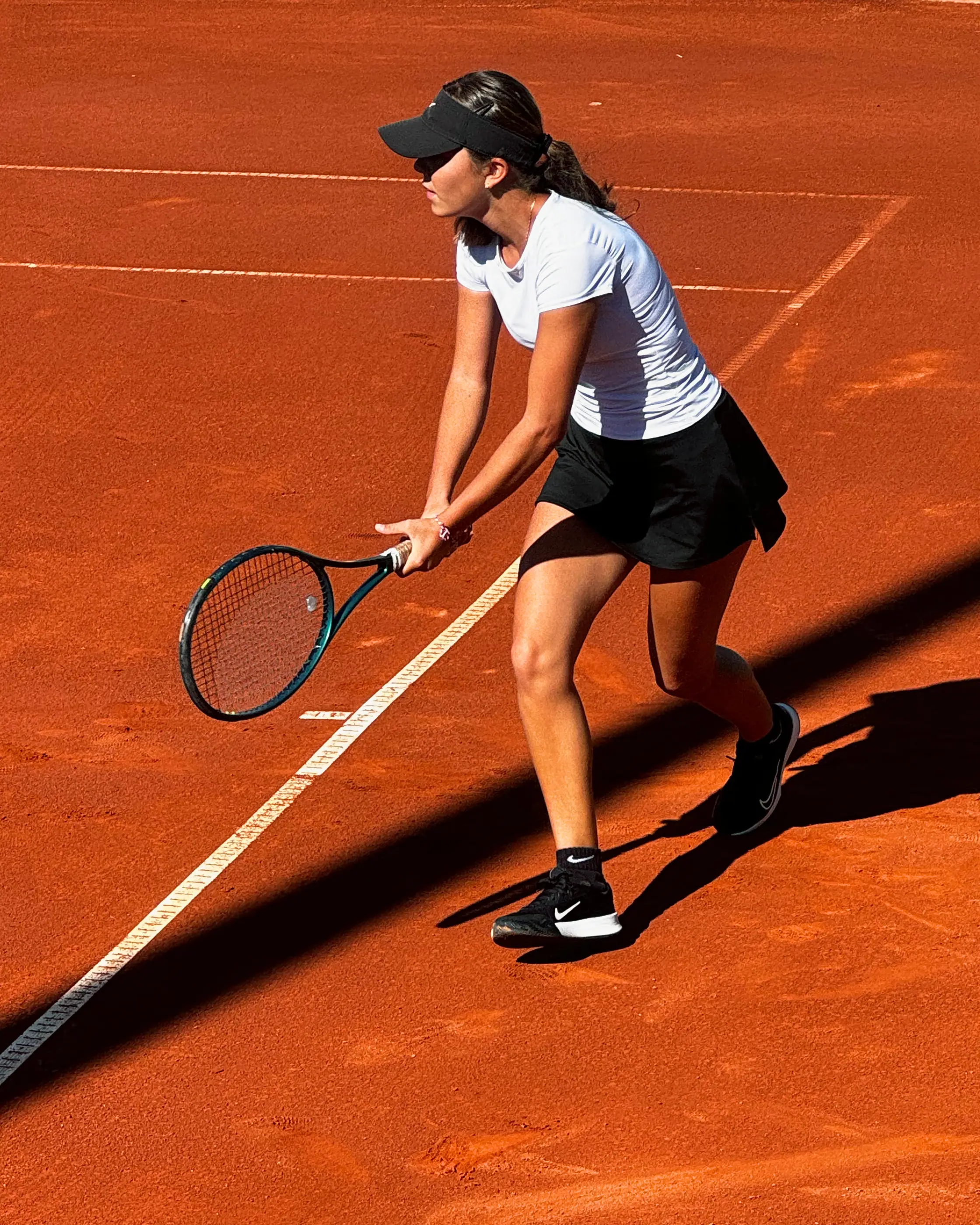 Nicole en posición de espera sobre una pista de tierra batida, vista desde arriba: rodillas flexionadas, la raqueta baja con las dos manos y una sombra larga cruzando la pista.