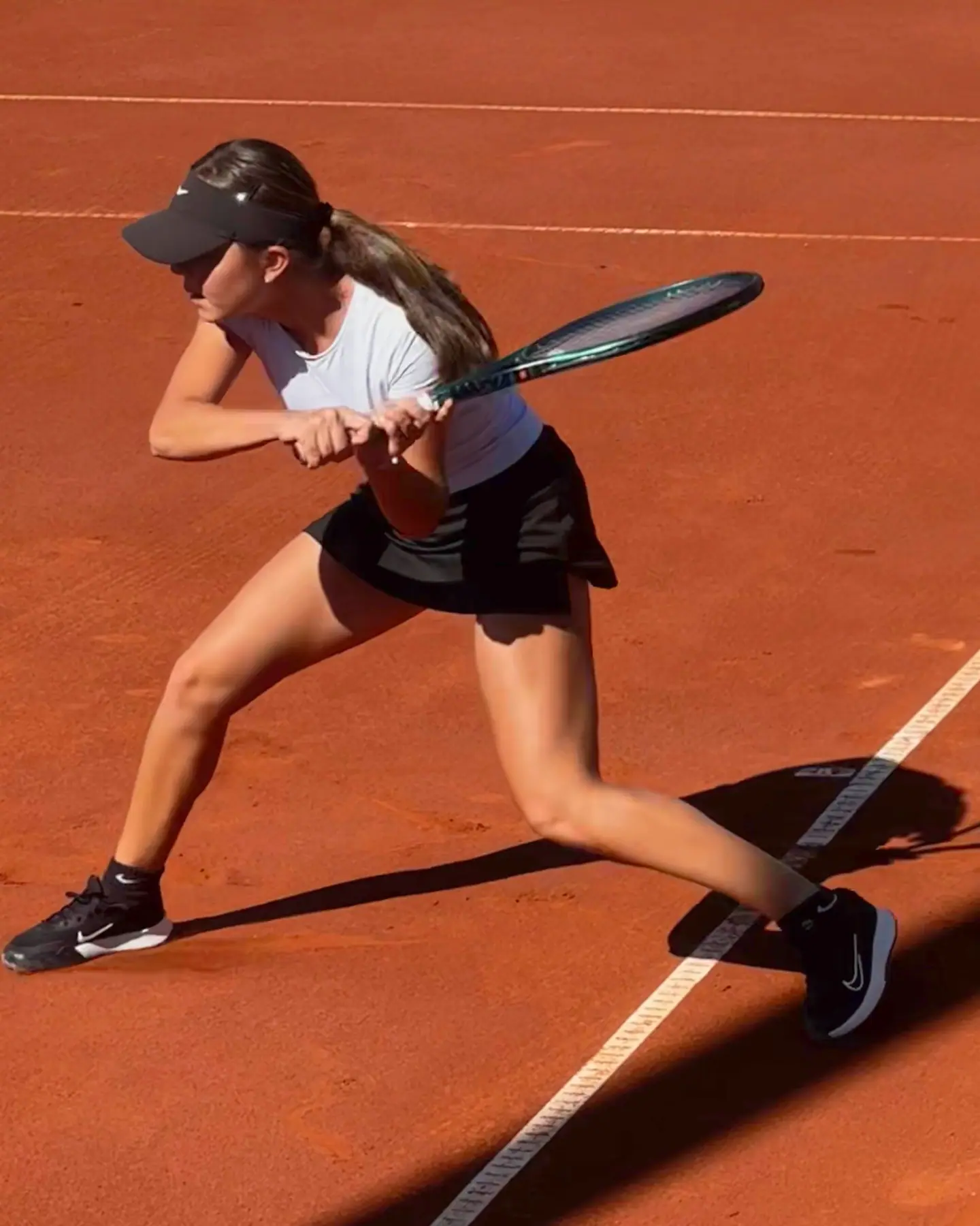 Nicole en un paso largo para un revés a dos manos en tierra batida, vista desde arriba, con la raqueta atrás y una sombra larga sobre la pista.