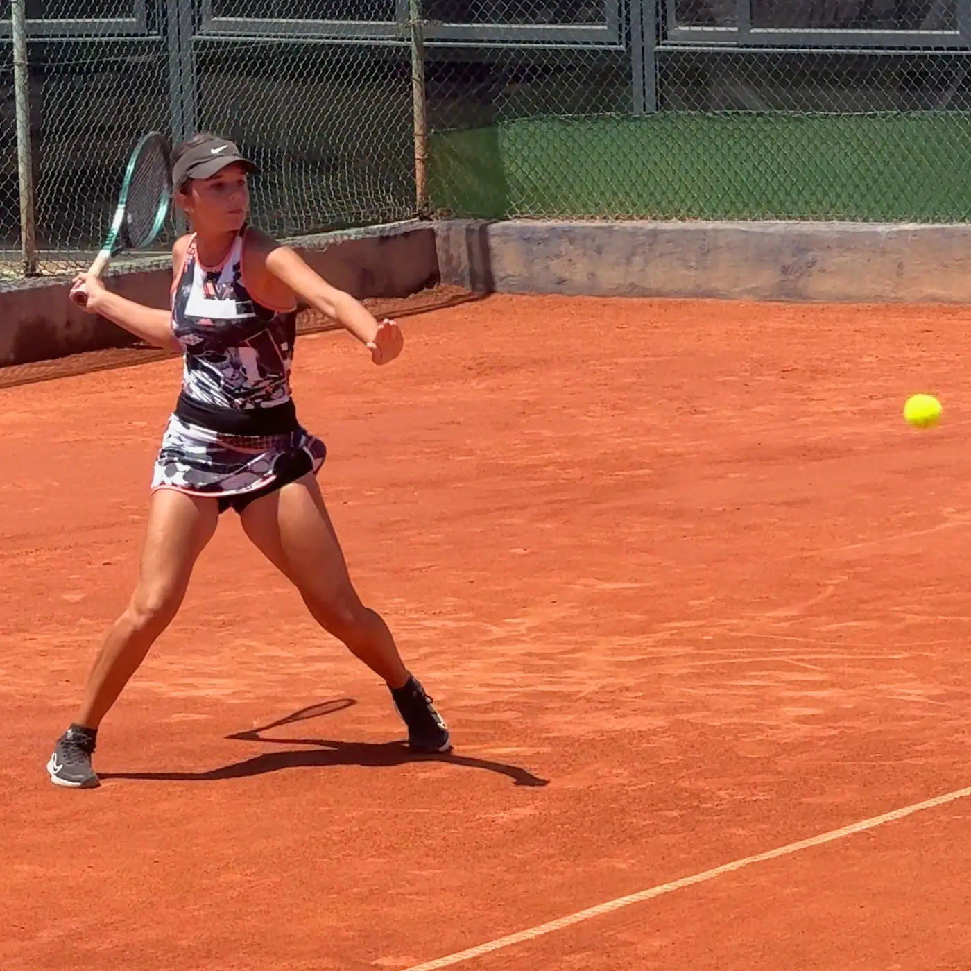 Nicole de frente a la cámara, con las piernas abiertas en una pista de tierra batida y la raqueta atrás para una derecha; la bola en el aire, a un lado.